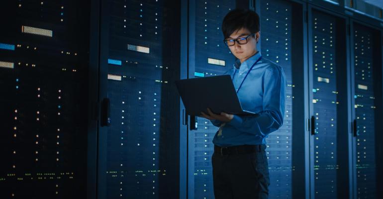 In a dark data center, a cloud security architect walks along a row of operational server racks.