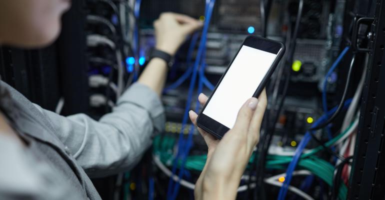 Close up of data center network technician holding smartphone with blank screen while connecting cables in outsourced server cabinet.